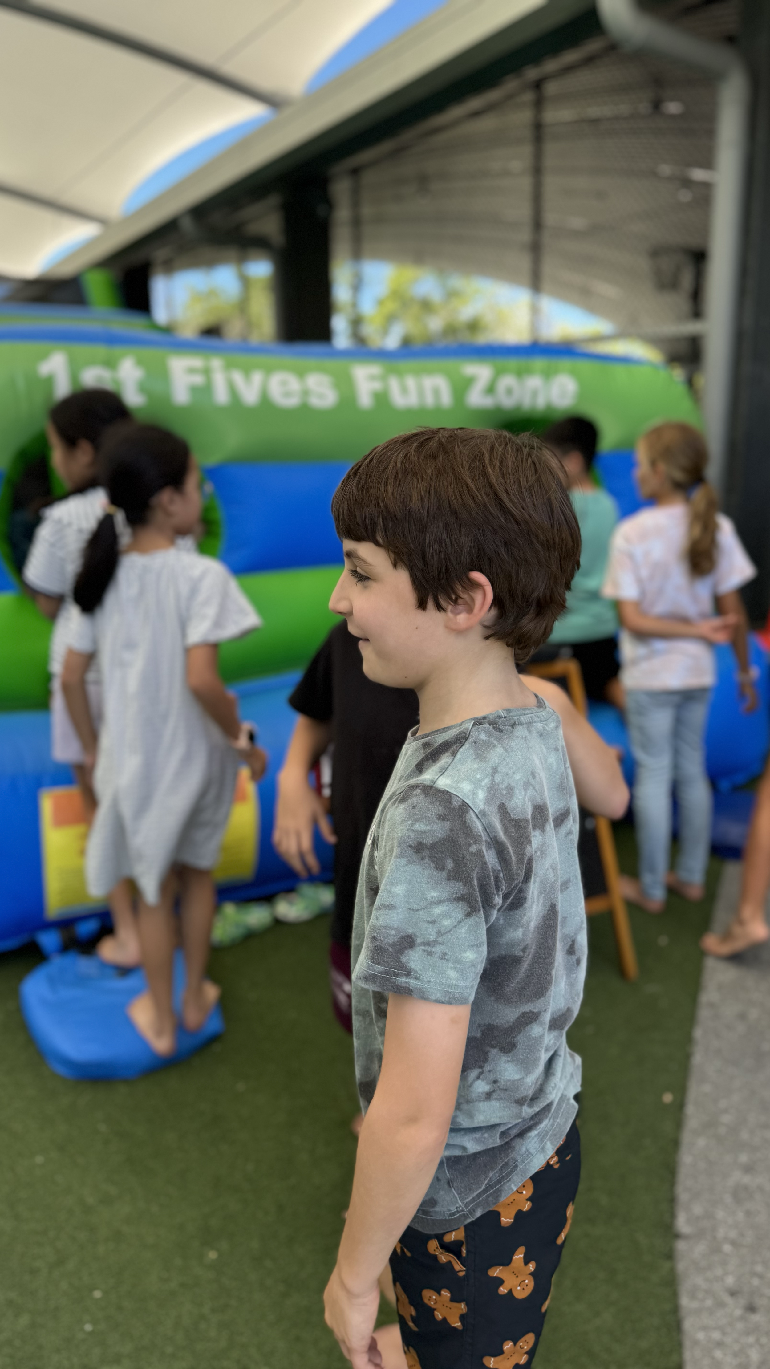 Boy standing in front of a bouncy house at a fun zone event, surrounded by children who are waiting in line.