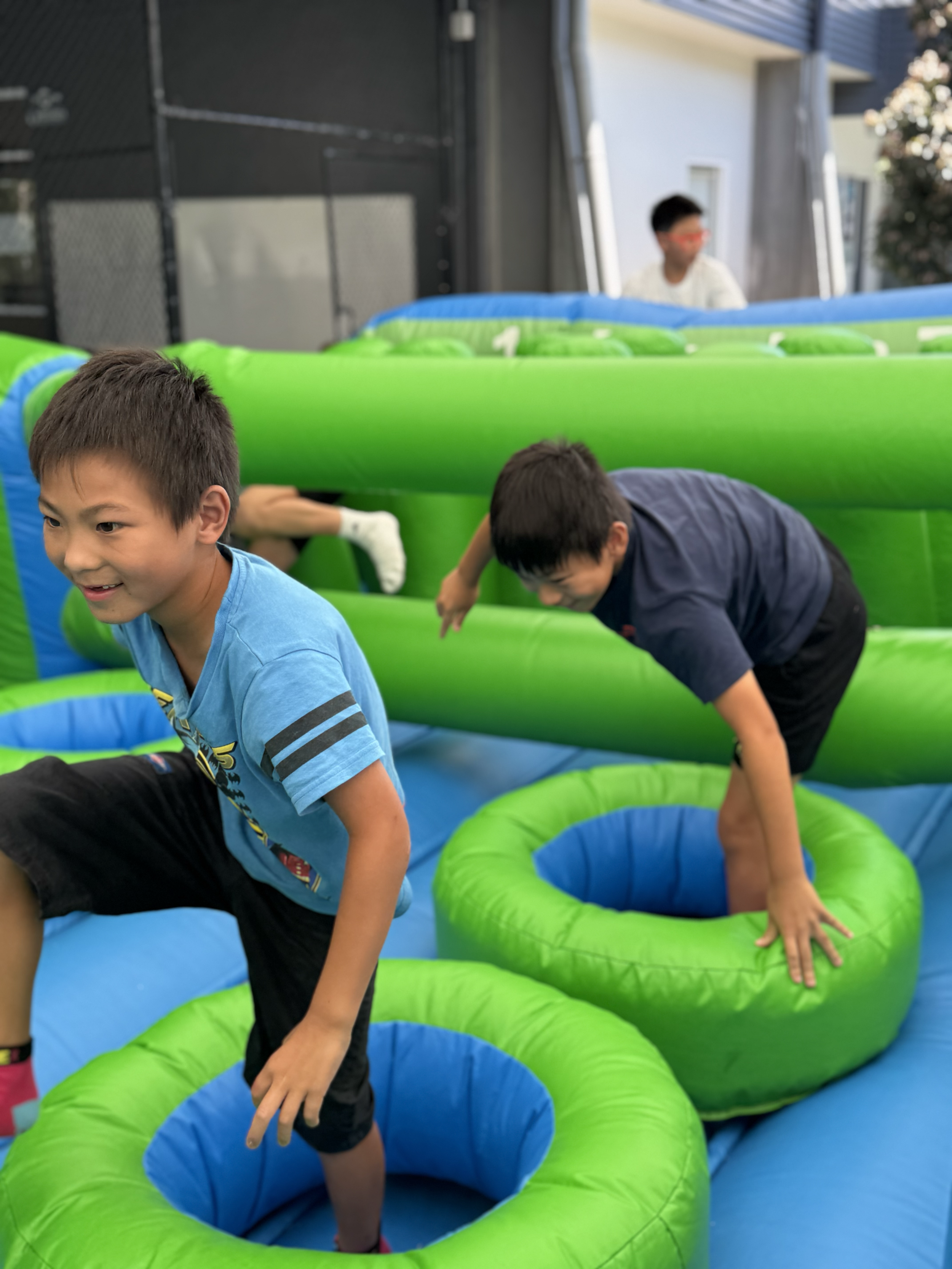 Children playing on an inflatable obstacle course outdoors with green and blue sections and a black fence in the background.