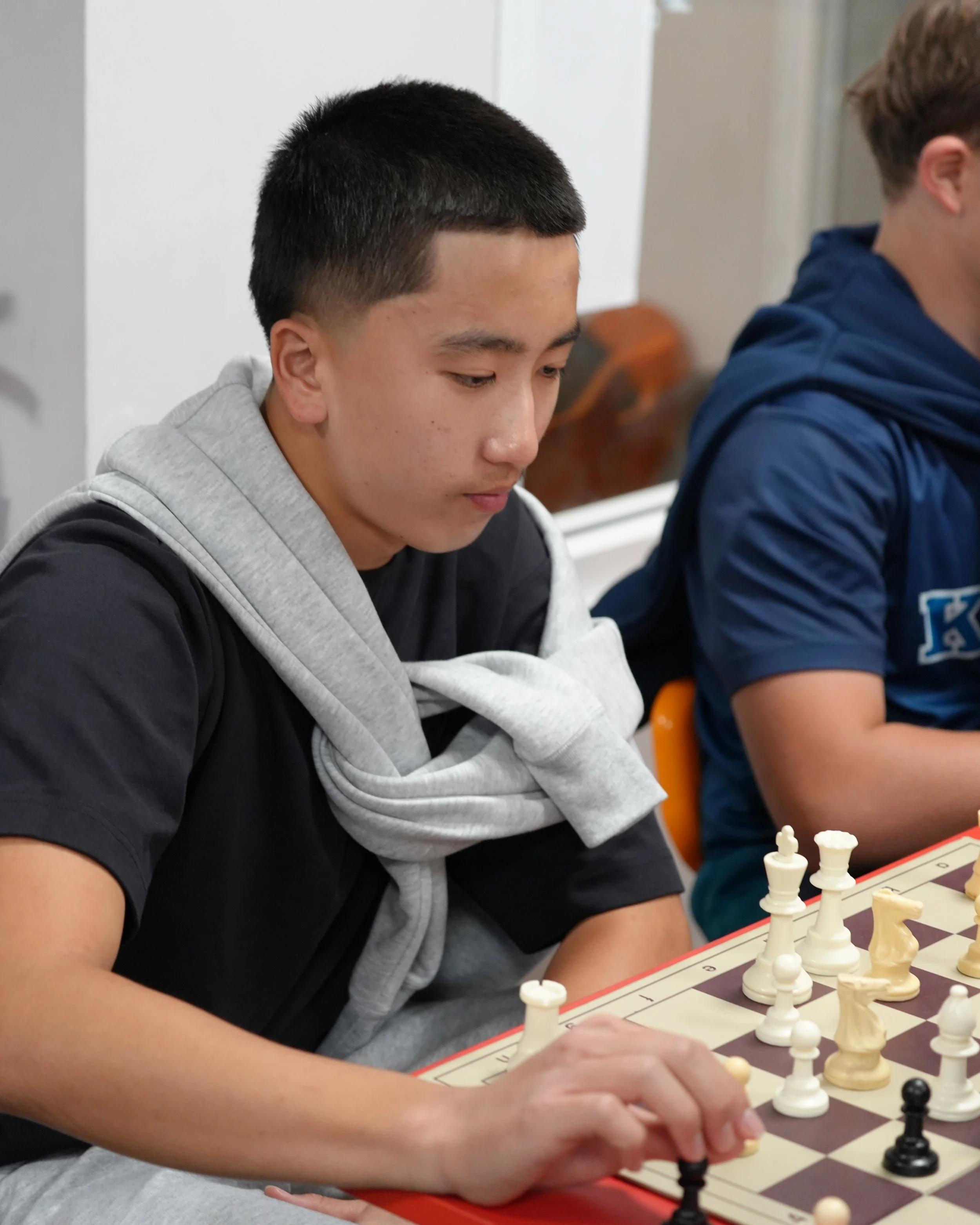 Two boys playing chess at a desk in a classroom, with other children in the background.