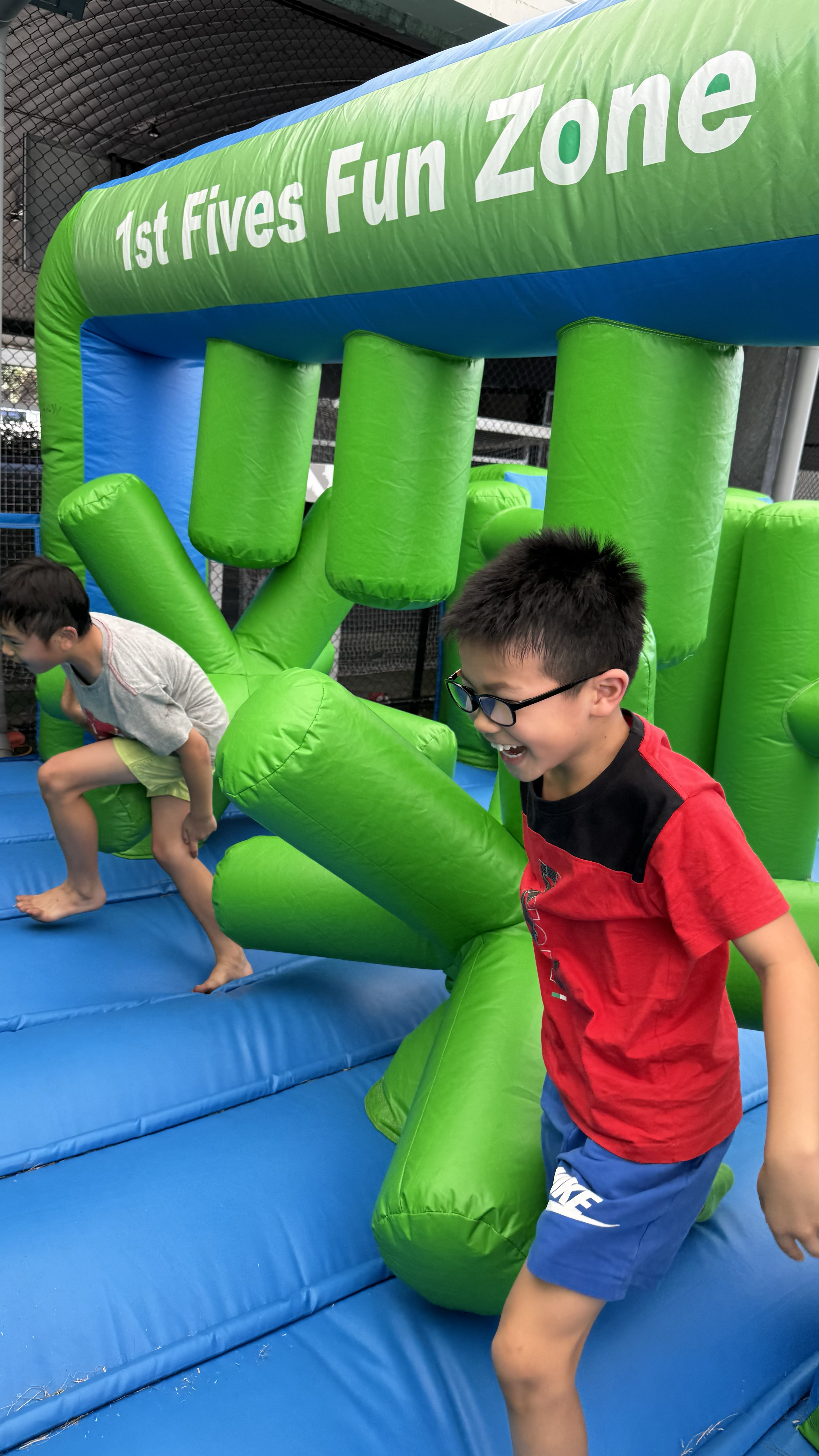 Two boys playing on an inflatable bounce house at a place called 1st Fives Fun Zone, with one boy climbing on it and the other laughing.