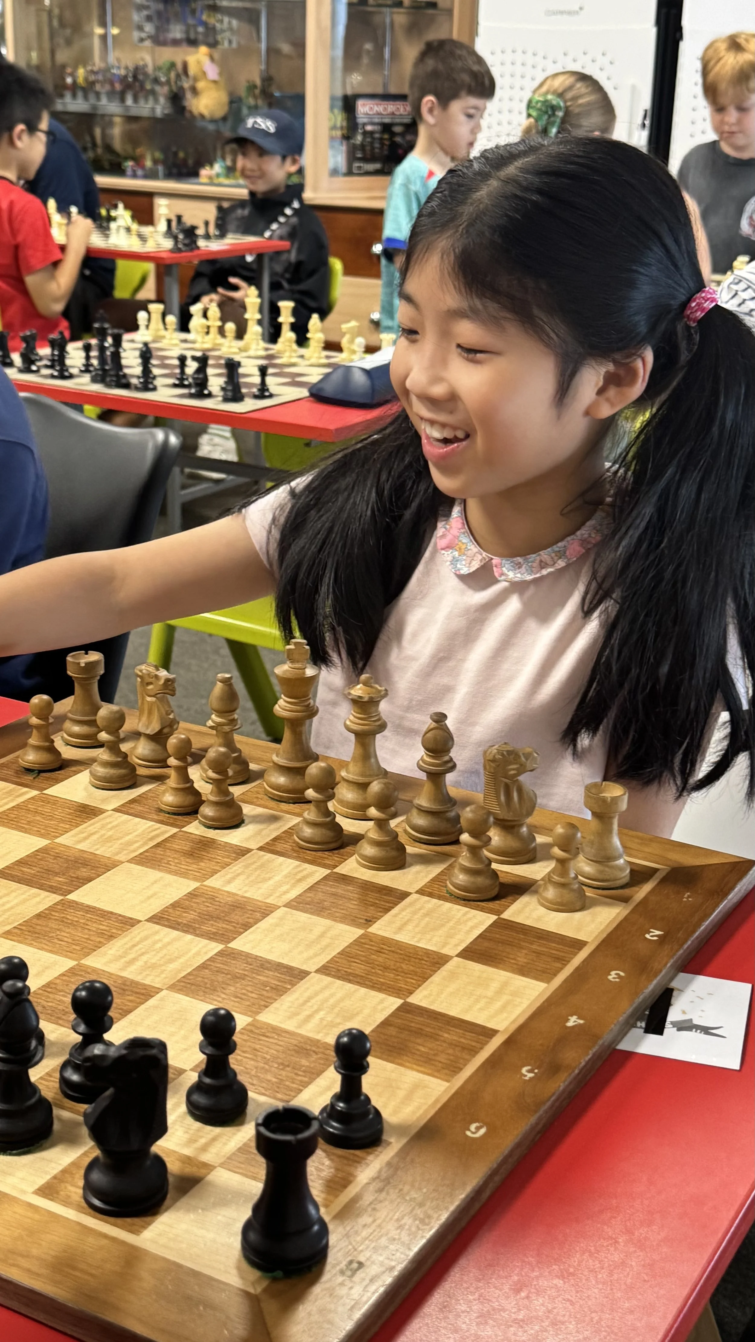 A young girl with long black hair in pigtails playing chess at a table in a busy game room with other children and chess sets visible in the background.