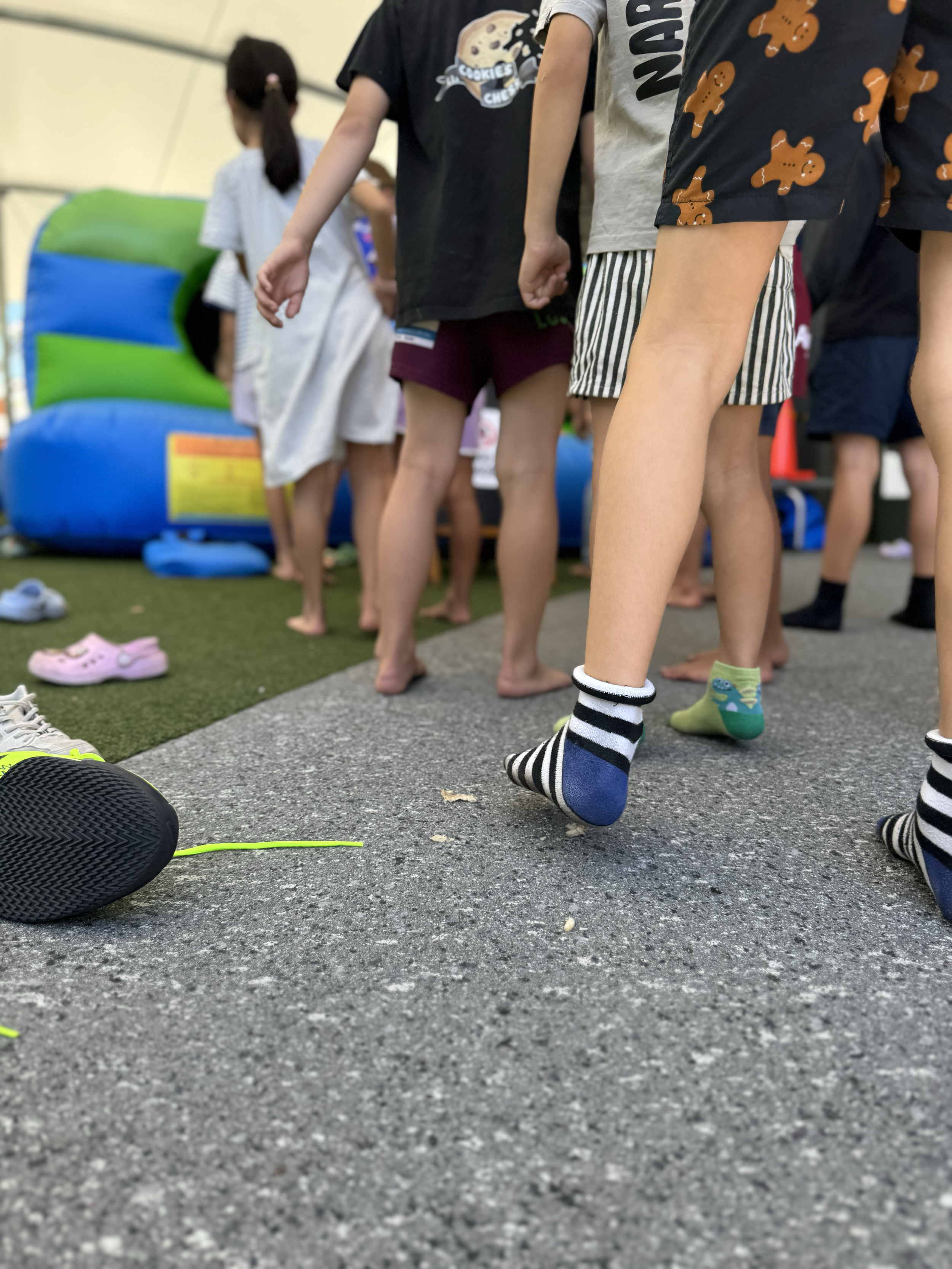 Children standing and waiting in line at an indoor playground, with some wearing socks and others barefoot, and a colorful inflatable slide visible in the background.