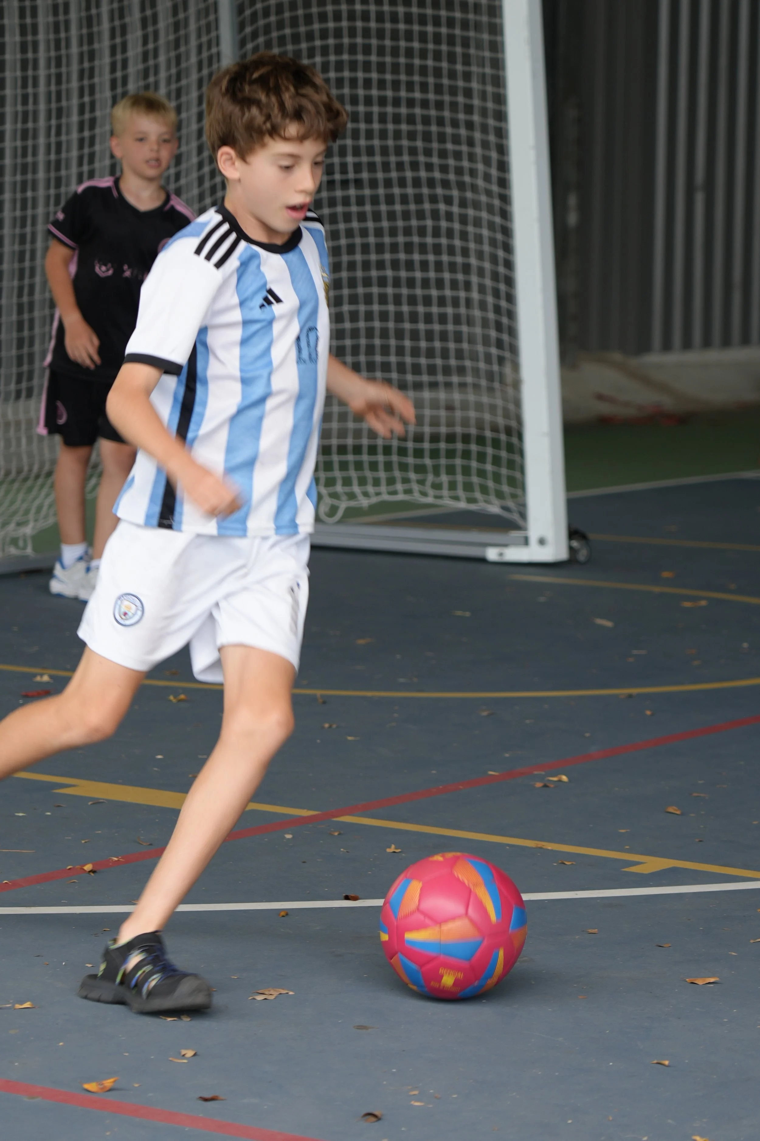 Two children playing soccer indoors, with one kicking a pink and blue ball and the other standing near a goal.
