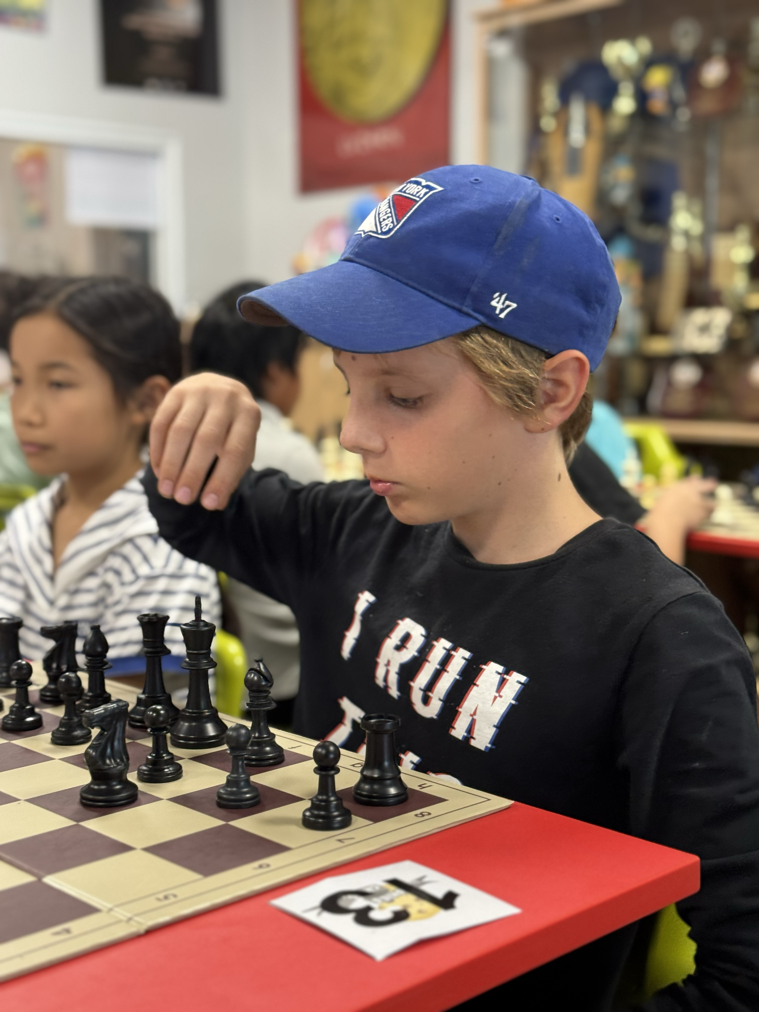 A young boy in a blue baseball cap and a black shirt playing chess in a lively indoor setting with other children in the background.