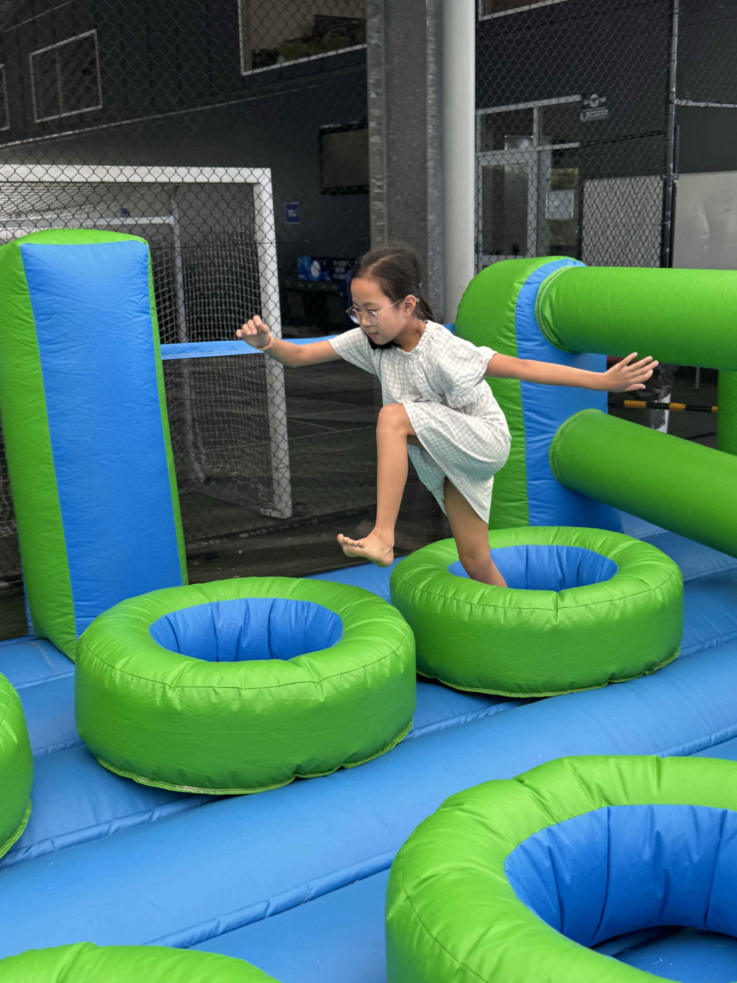 A girl in a light-colored dress balancing on an inflatable obstacle course with a circular hole, surrounded by colorful padded barriers and tubes, set in an outdoor play area.