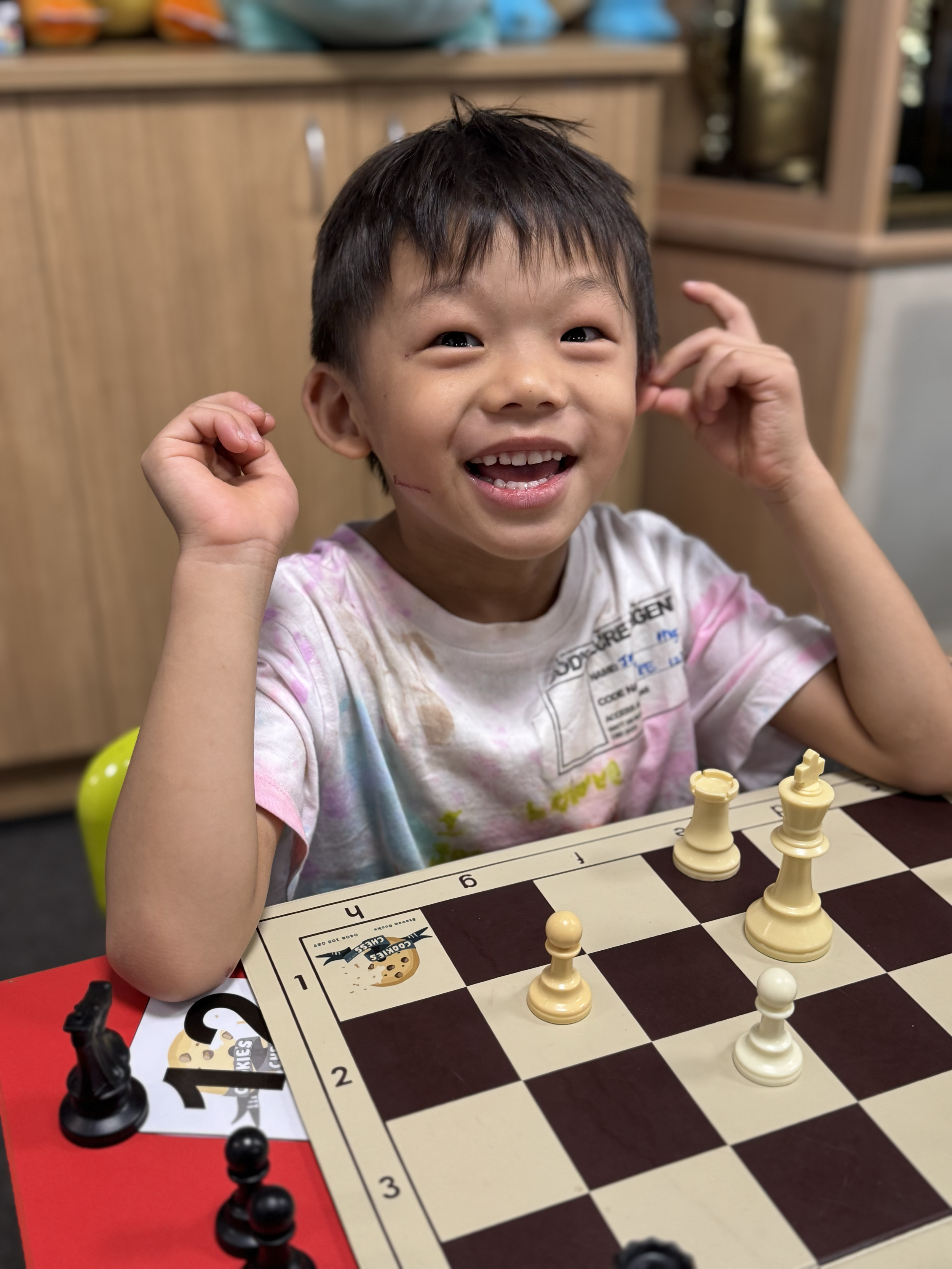 A young boy with short dark hair, smiling excitedly, sitting at a chessboard with several chess pieces, including pawns, rooks, a bishop, and a king, on the board, in a room with wooden cabinets in the background.