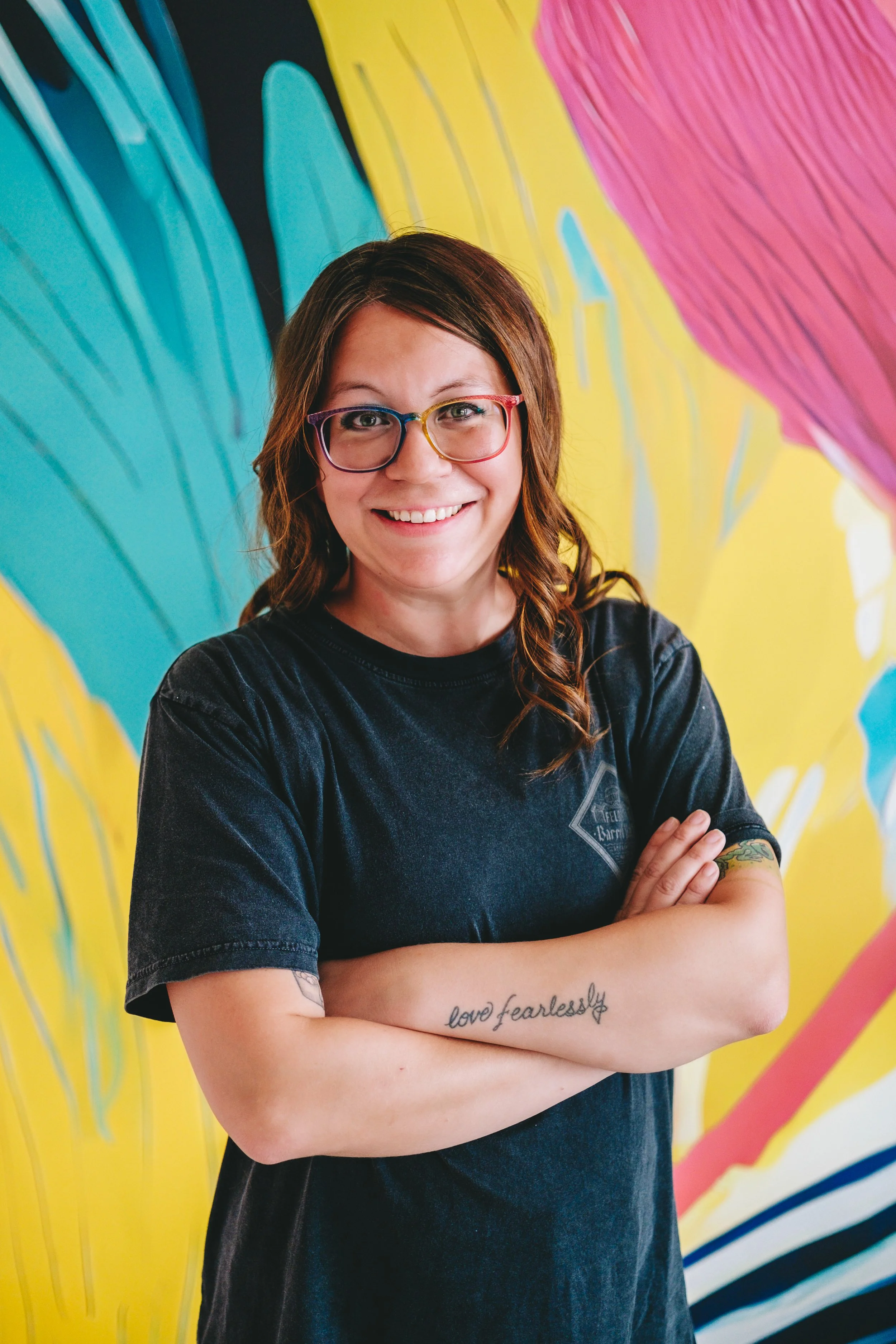 A smiling woman with shoulder-length brown hair, wearing rainbow-colored glasses and a black T-shirt, stands with arms crossed in front of a colorful mural featuring large, abstract shapes in blue, pink, yellow, and black.
