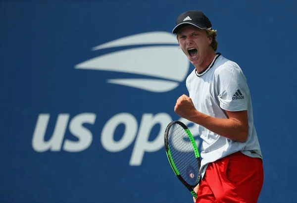 Tennis player celebrating in front of the US Open logo during a match, holding a racket and wearing a cap, white shirt, and red shorts.