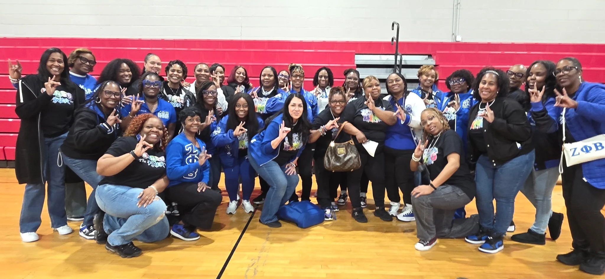 A group of women posing together in a gymnasium, many making the 'Lambda' hand sign, wearing blue and black jackets and shirts, indoor gym with red bleachers in the background.