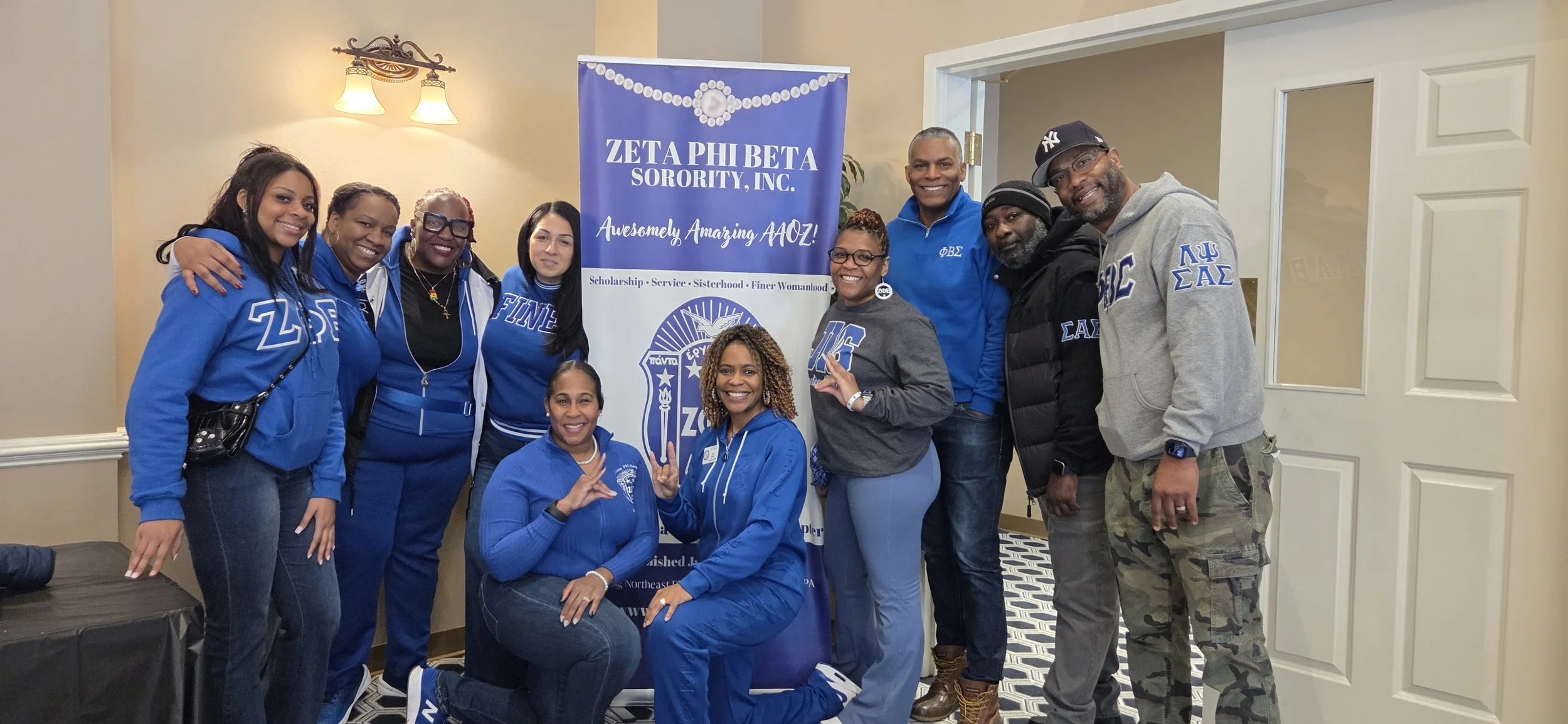 A group of ten people, mostly women, standing and kneeling in front of a blue banner for Zeta Phi Beta Sorority, Inc. They are indoors, smiling, and dressed in casual blue and gray clothing, some with sorority logos or symbols.