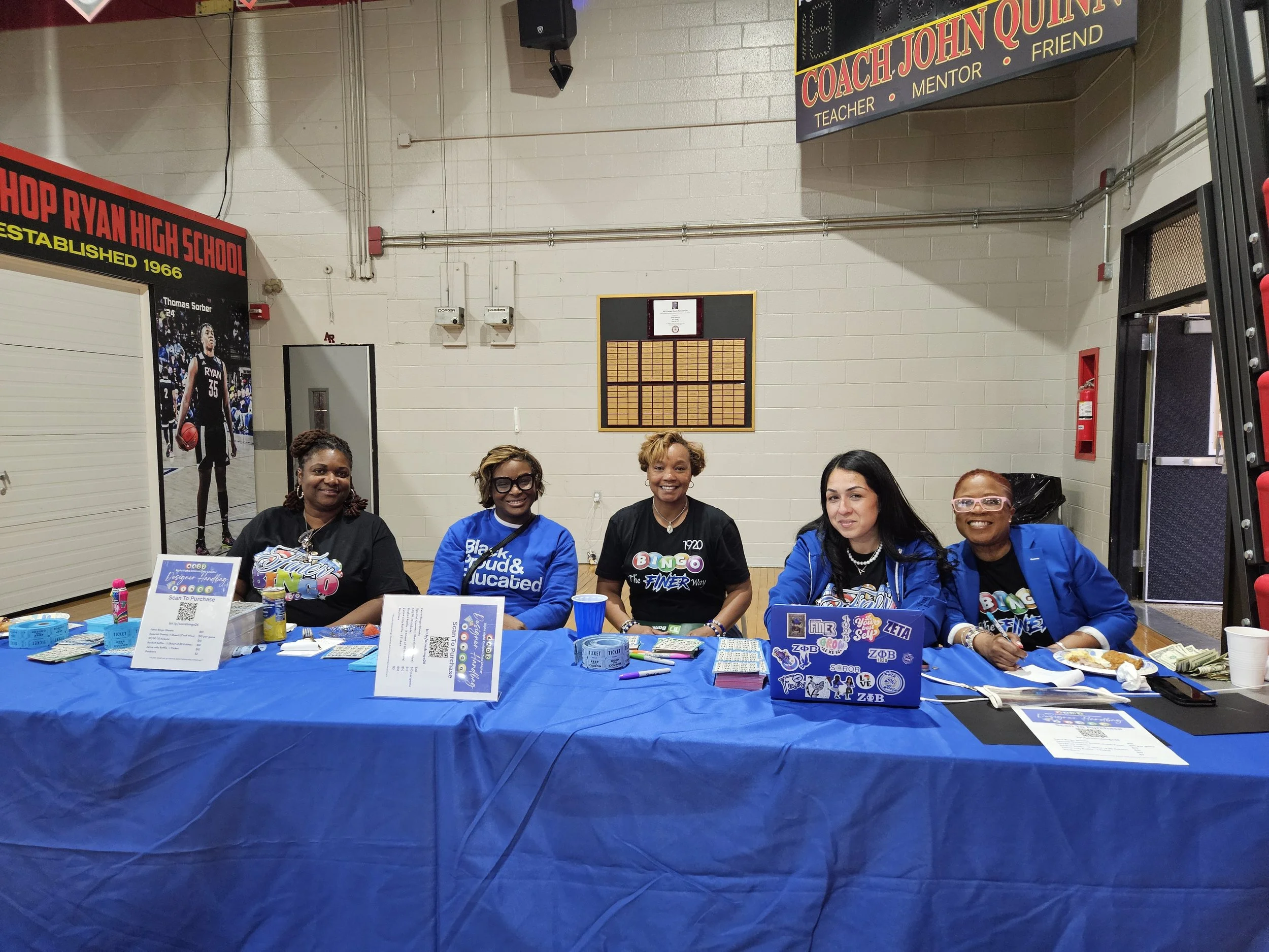 Group of women sitting behind a table covered with a blue tablecloth, displaying promotional materials and raffle tickets, inside a gymnasium. Walls and posters are visible in the background.