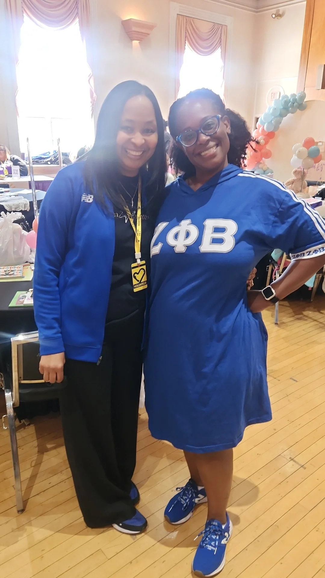 Two women smiling and standing together inside a decorated room. One woman is wearing a blue jacket and the other is wearing a blue dress with Greek letters. Balloons and tables are visible in the background.