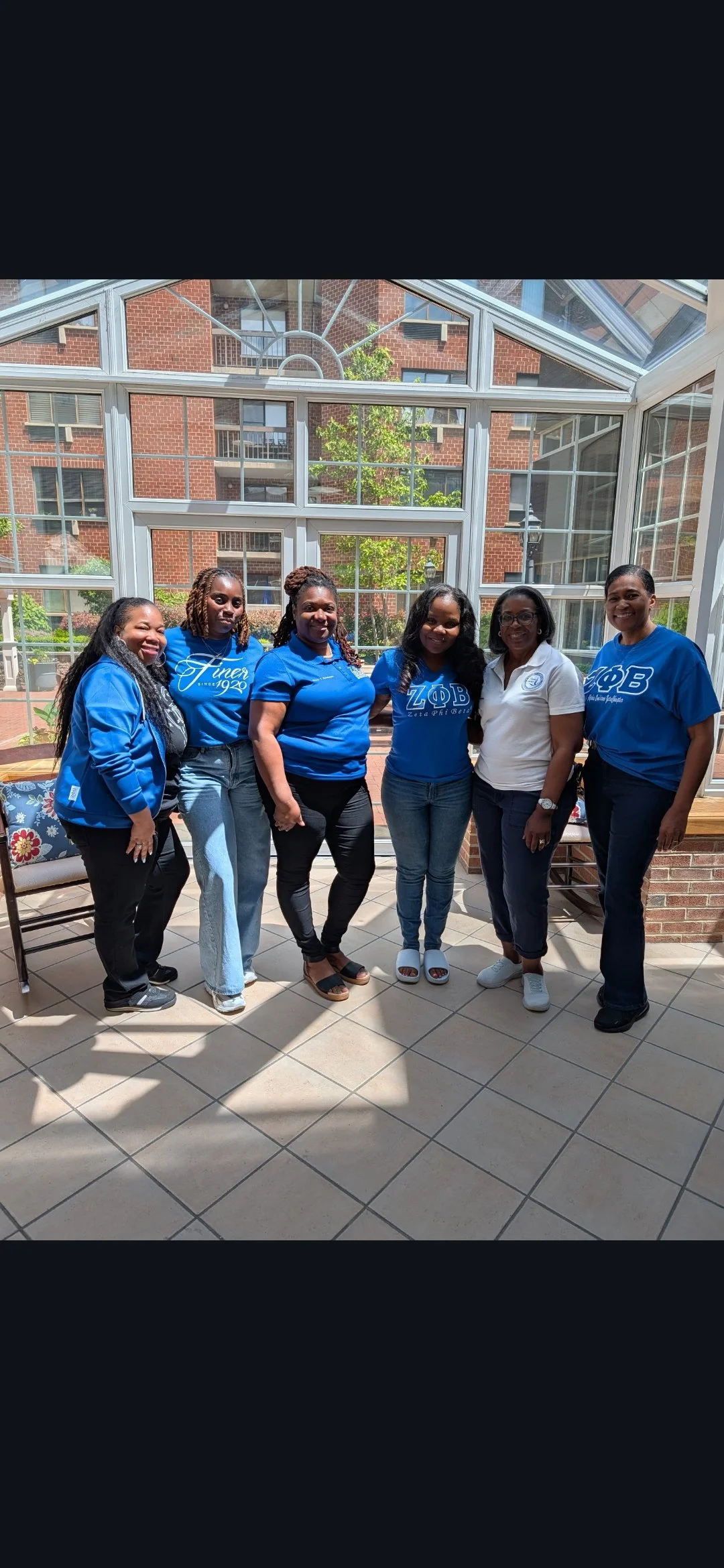Six women standing inside a sunlit glass enclosure, wearing blue shirts with Greek letters and one woman in white, with a brick building and trees visible outside.