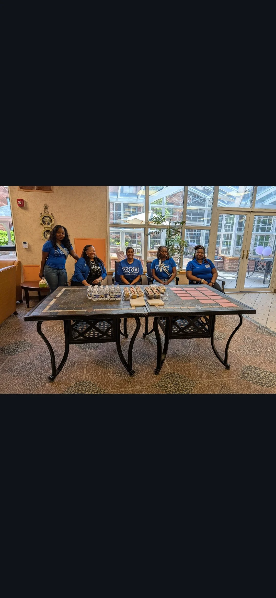 Five women standing and sitting behind a table with water bottles, cupcakes, and cookies in a bright indoor space with large windows and glass doors.