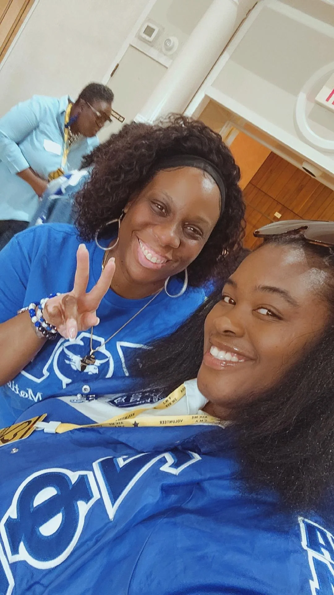 A woman taking a selfie with an older woman wearing a blue shirt, both smiling and making a peace sign. They are in a room that appears to be a hospital or medical facility.