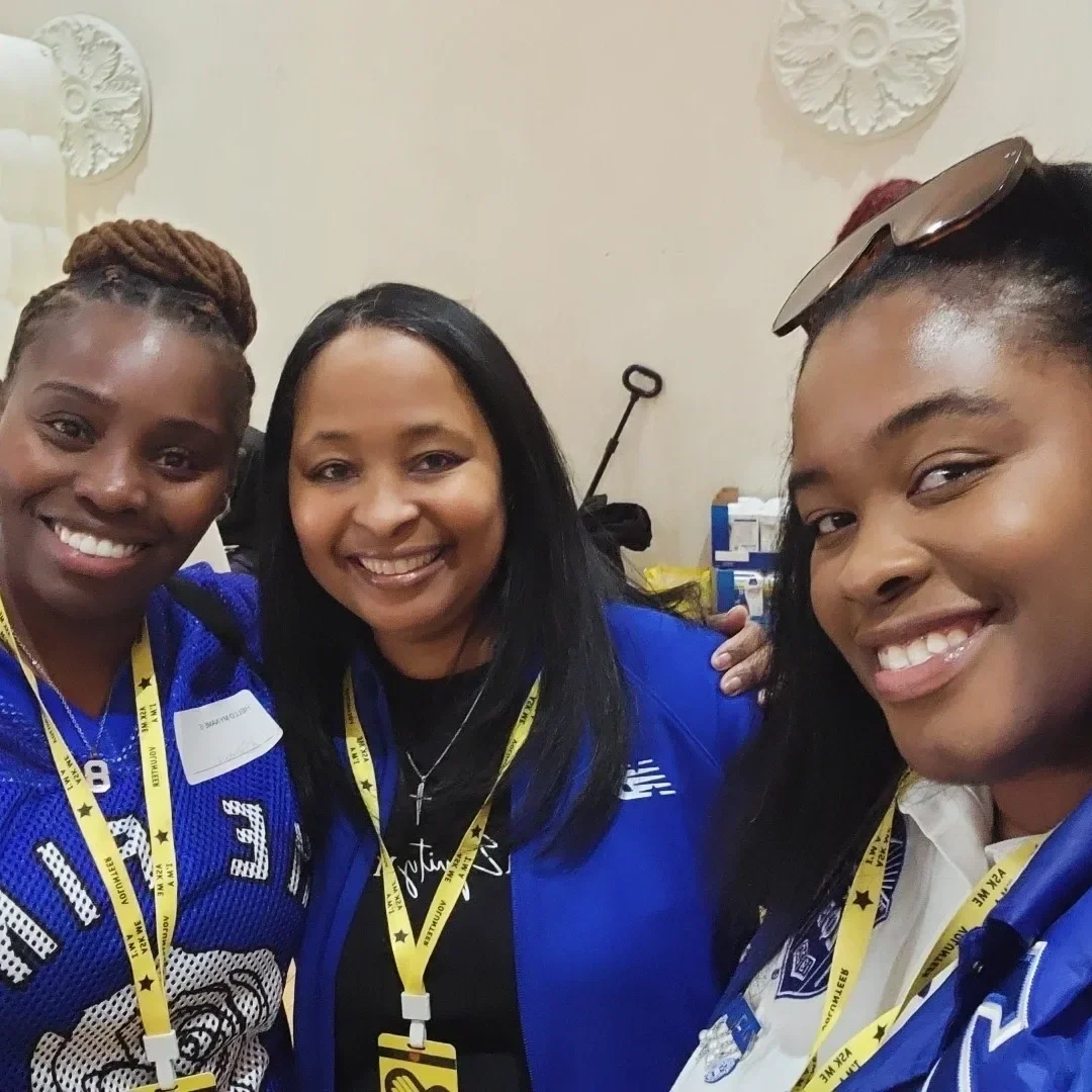 Three women smiling for a selfie, all wearing blue sports jerseys and yellow lanyards, in a room with decorative wall hangings.