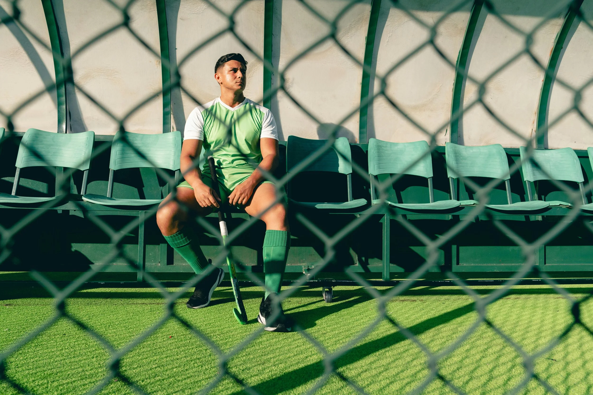 Athlete sitting alone on a bench behind a chain-link fence holding a hockey stick — illustrating the isolation and pressure athletes face beyond the field