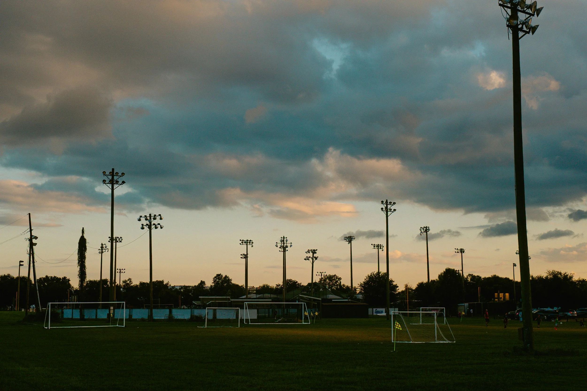 Empty soccer field at sunset with stadium lights and dramatic sky — representing the quiet weight of sports culture and athlete mental health
