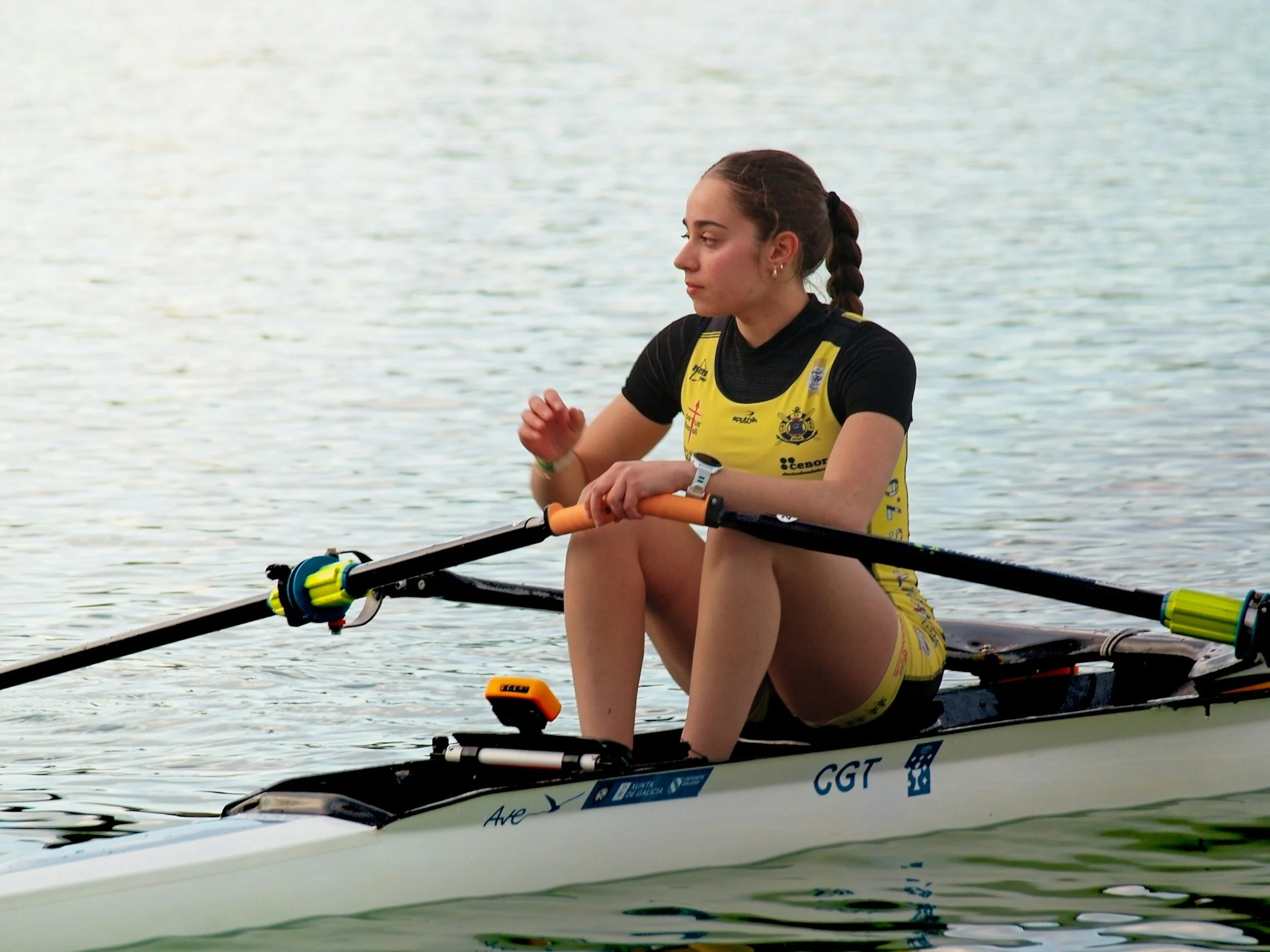 Young female athlete pausing in a rowing shell on calm water — reflecting the mental and emotional demands behind athletic performance