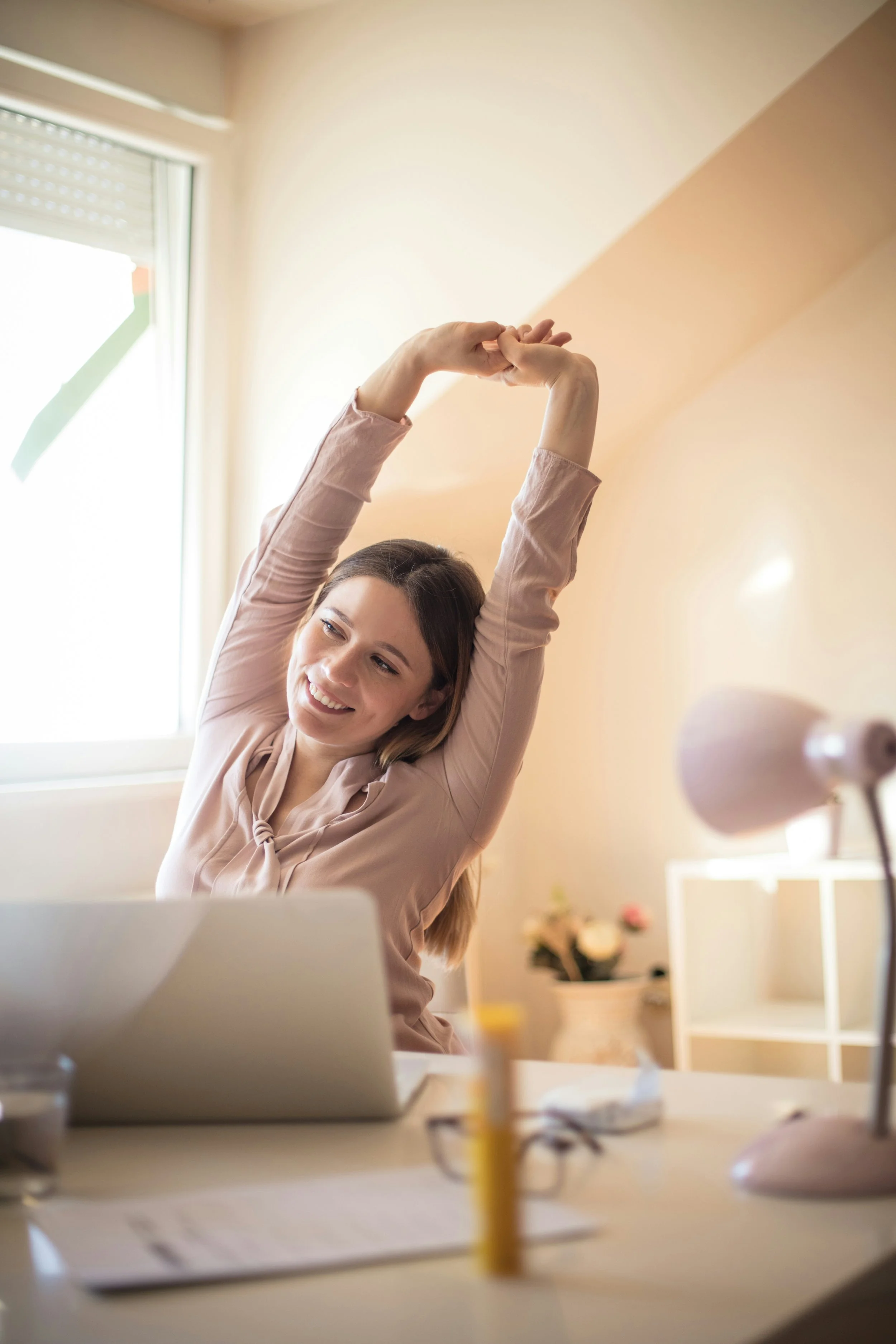 A brunette female is stretching at her desk happily. She is taking an important moment for mindfulness and movement during her busy day as a work from home professional.