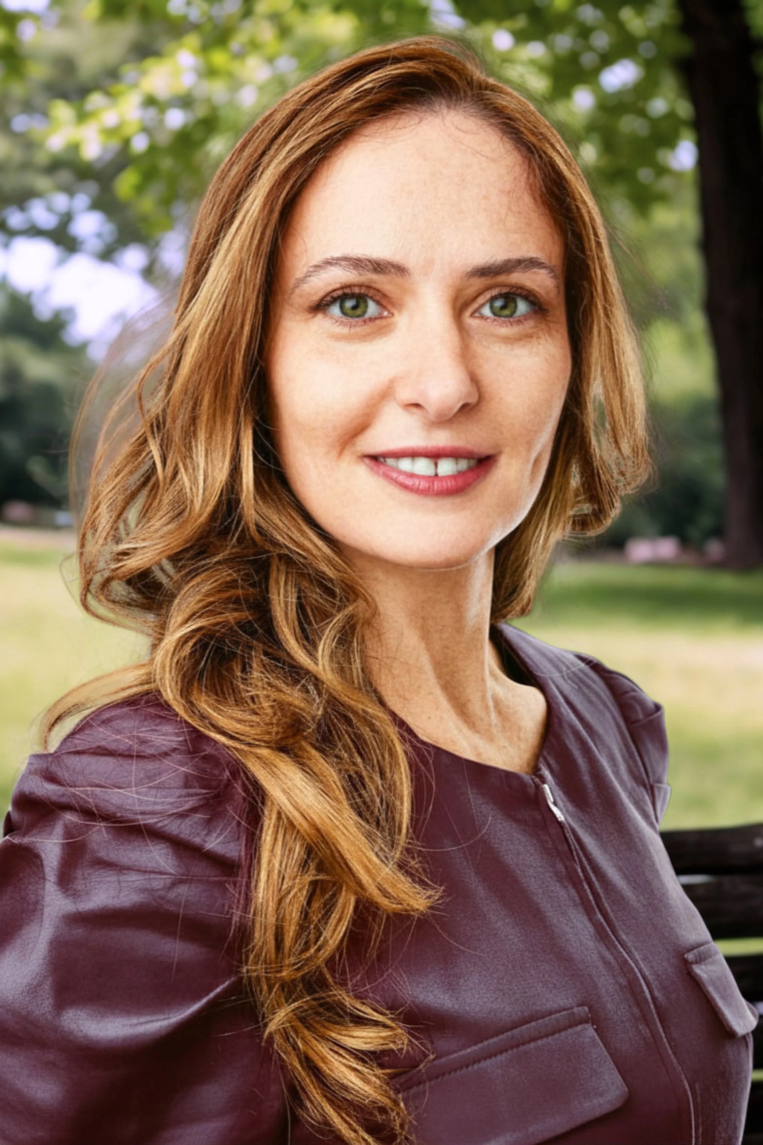 A woman with long, wavy red hair and green eyes smiling outdoors in a park setting.