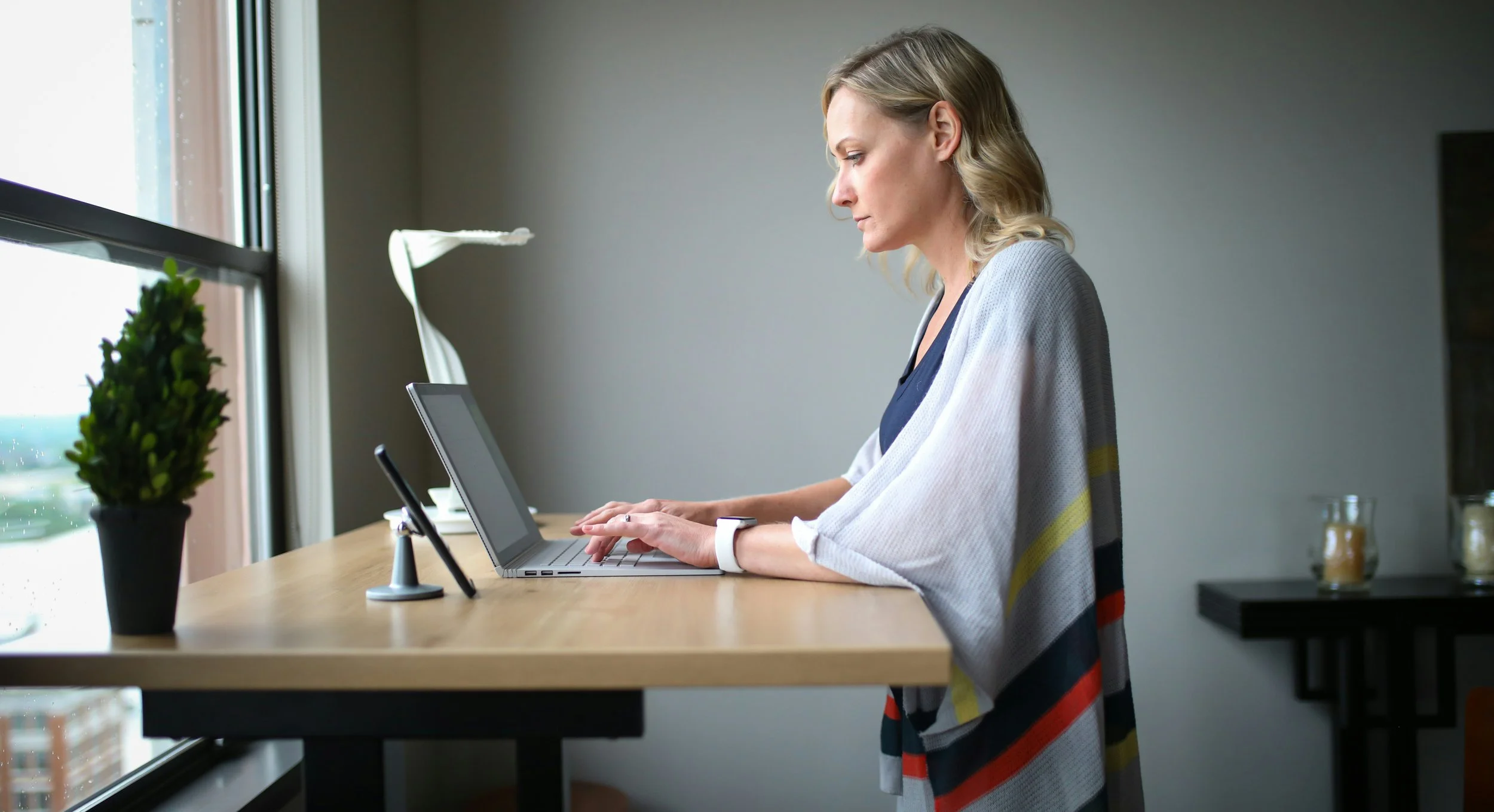 A blonde professional woman working at a standing desk, looking focused but strained, illustrating the concept of "quietly cracking" and high-functioning anxiety in the workplace.