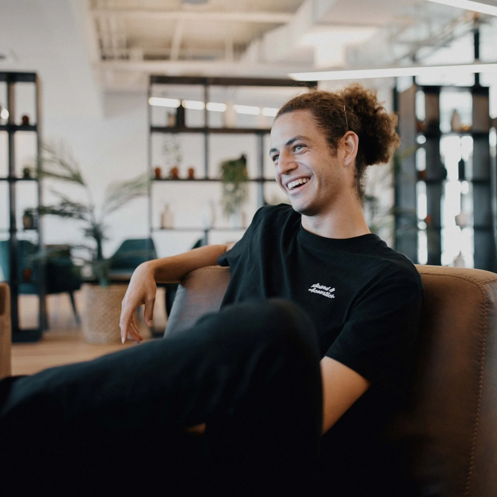 Young man with curly hair smiling and sitting relaxed on a brown couch in a modern, well-lit office space with plants and shelving in the background.