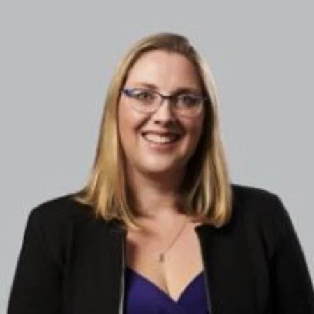 A woman with blonde hair wearing glasses, a black blazer, and a blue top, smiling at the camera.