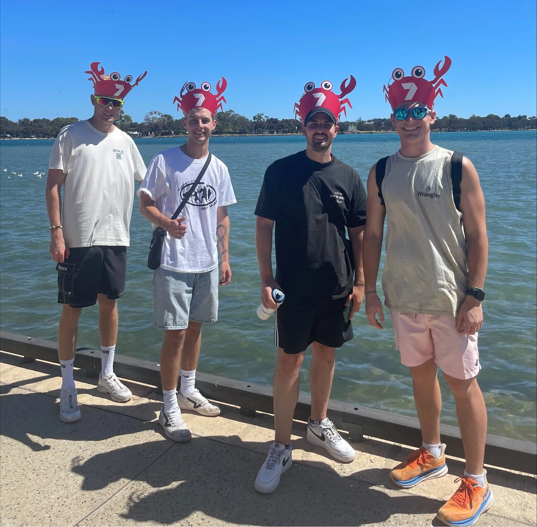 Four young men standing by a lake wearing crab themed hats with the number 7 on them. They are dressed casually in summer clothes and sunglasses, with one holding a water bottle.