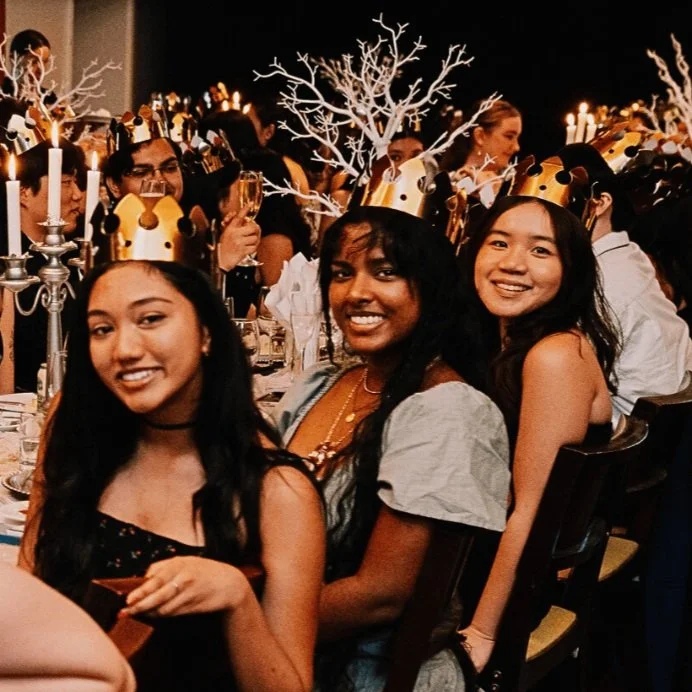 Group of young women celebrating at a party, wearing gold crowns, with decorative white branches and candles on the tables in the background.