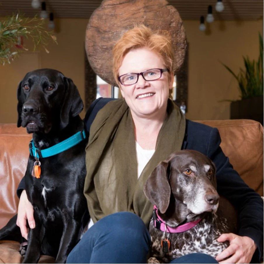 A woman with glasses and short red hair sitting on a brown leather couch with two dogs, one black with a blue collar and one brown and white with a pink collar, in a cozy indoor setting.
