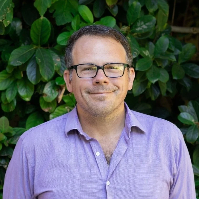 A man with glasses and short dark hair, smiling, wearing a light purple button-up shirt, standing in front of green leafy bushes.