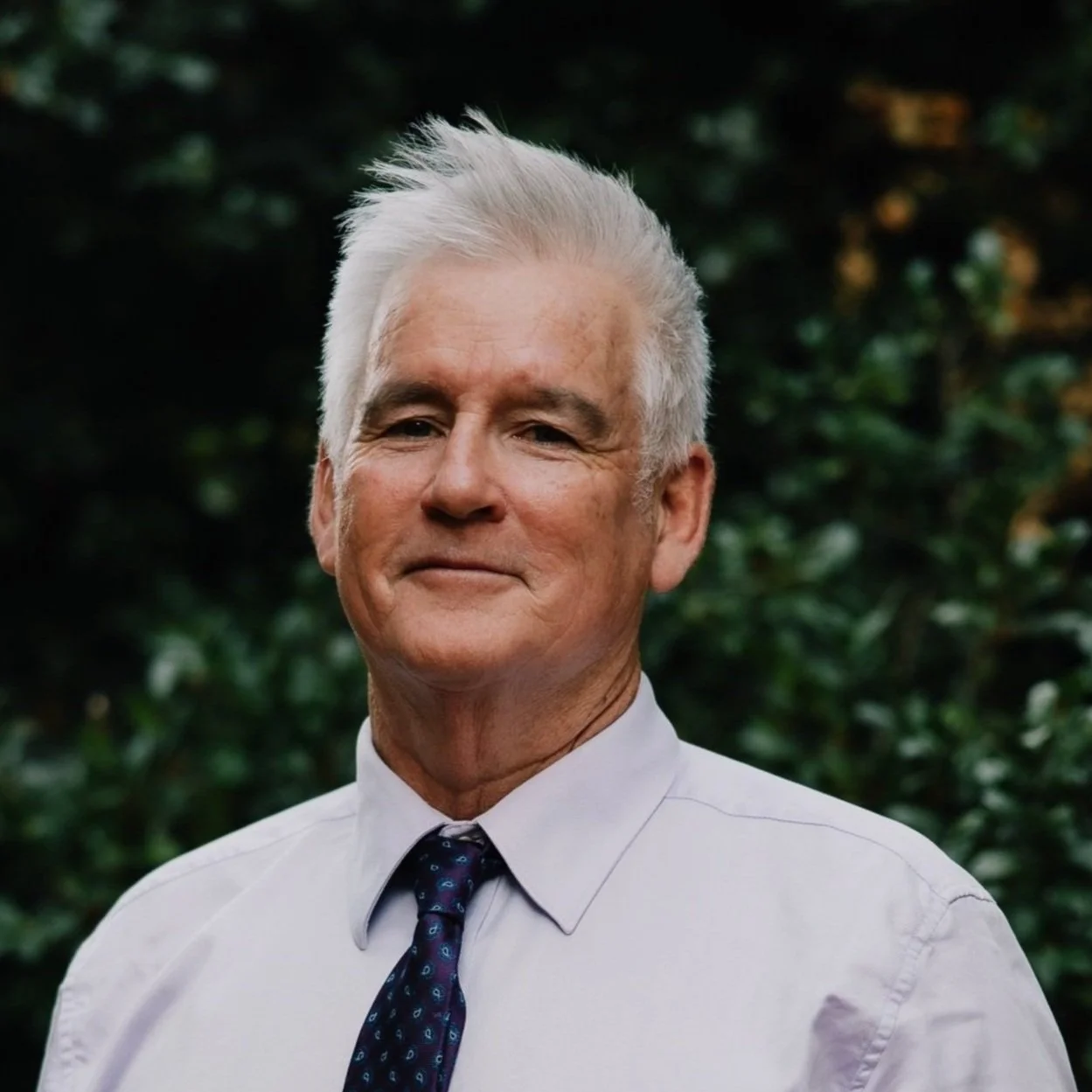 An elderly man with white hair wearing a light-colored shirt and a dark patterned tie, standing outdoors with a blurred green leafy background.