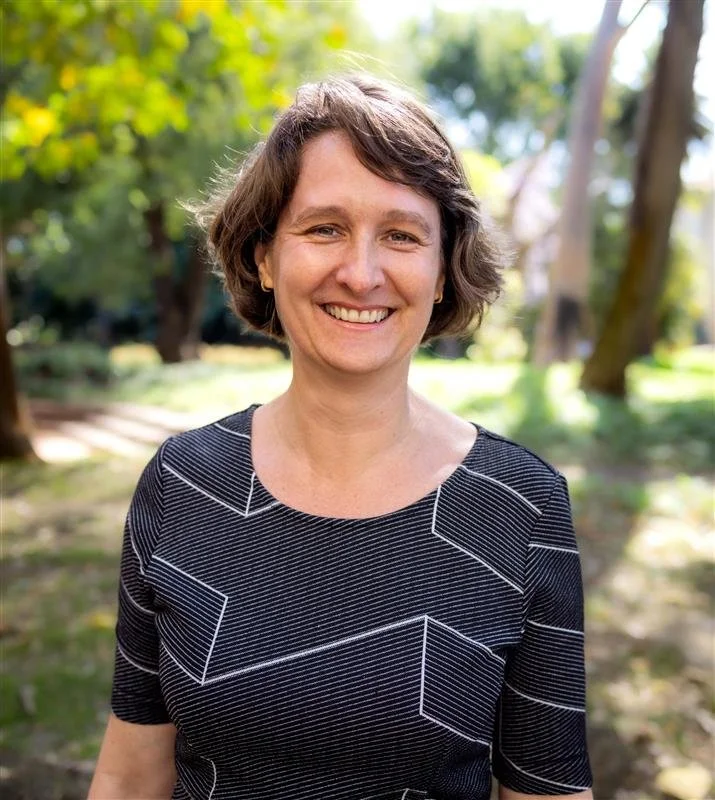 A woman with short brown hair smiling outdoors in a park with green trees in the background.