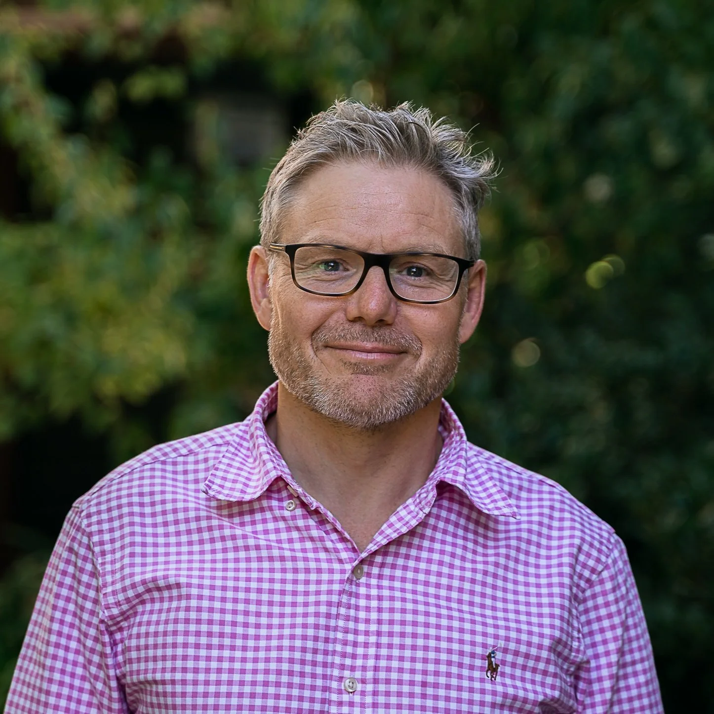 Close-up of a middle-aged man with gray hair and beard, wearing glasses and a pink checkered shirt, standing outdoors with green foliage in the background.