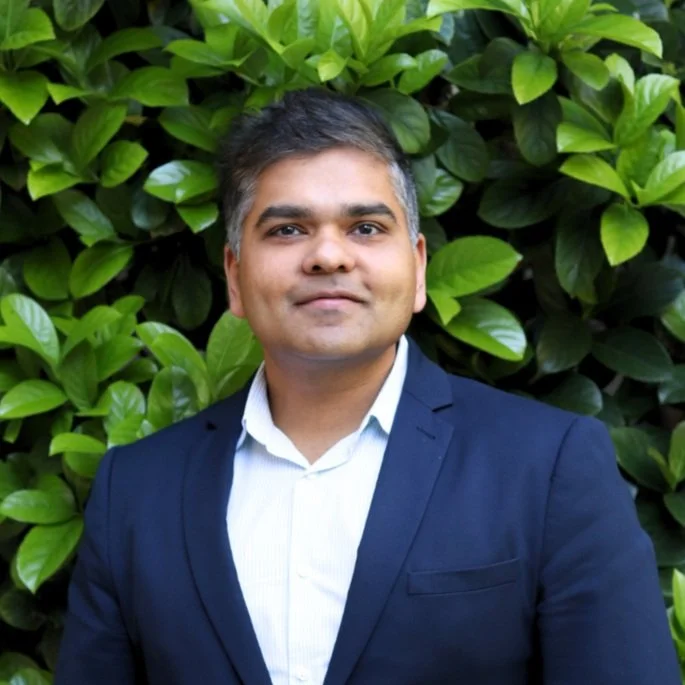 Man in suit standing outdoors in front of green leafy plants.