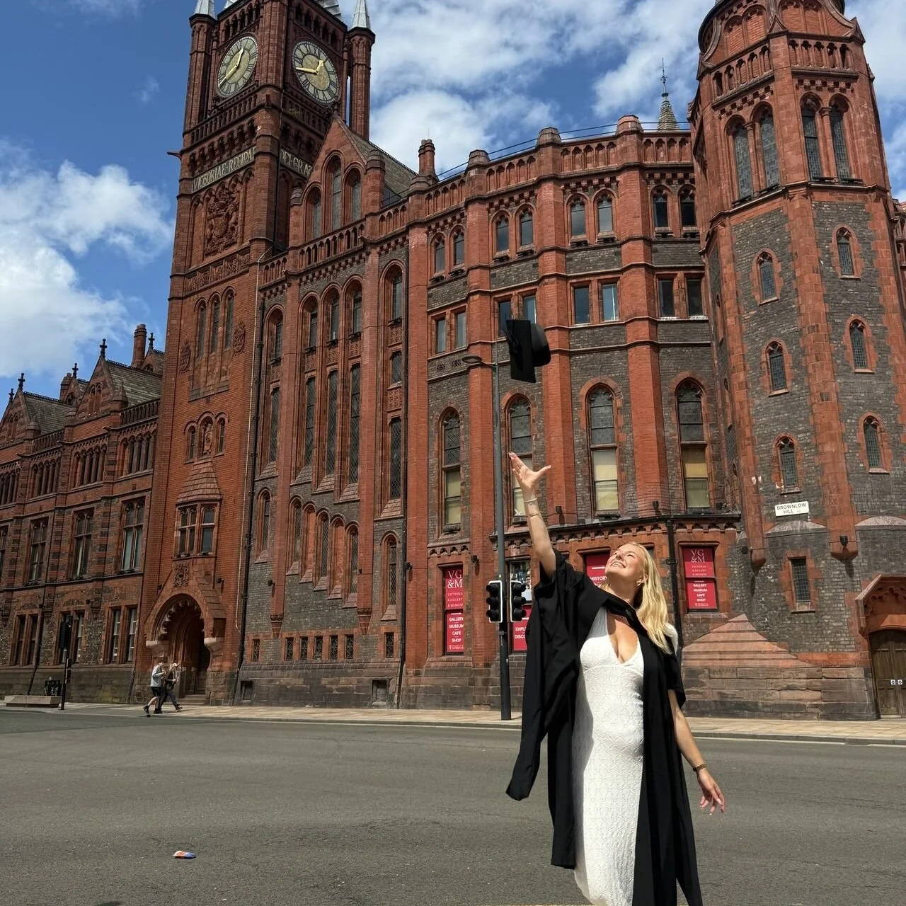 A woman in a white dress and black gown tossing a graduation cap into the air in front of a large, historic red-brick building with a clock tower, under a partly cloudy sky.