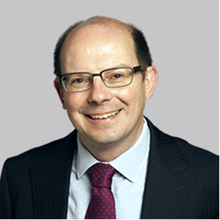 A man with glasses in a business suit, smiling against a gray background.