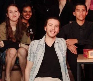 Group photo of five young adults, four sitting and one standing, smiling and looking at the camera in a casual setting.