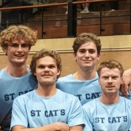 Group of young men in light blue T-shirts with 'ST CAT'S' printed on them, standing together in an indoor setting with wooden walls and a balcony.