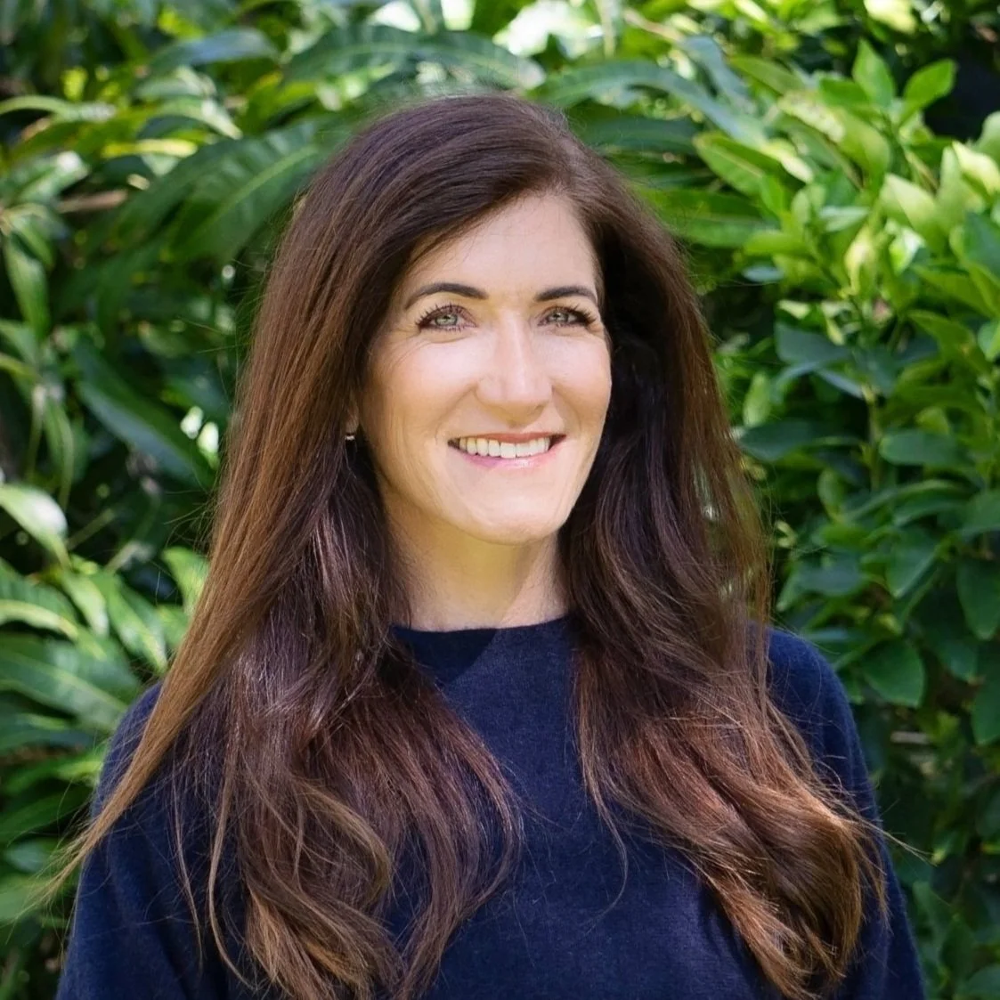 A woman with long brown hair smiling outdoors with green foliage in the background.