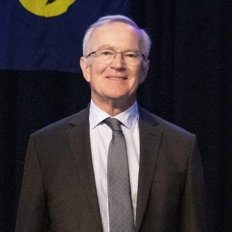 A smiling older man wearing glasses, a dark suit, a white striped shirt, and a gray tie, standing in front of a dark background with part of a flag visible.