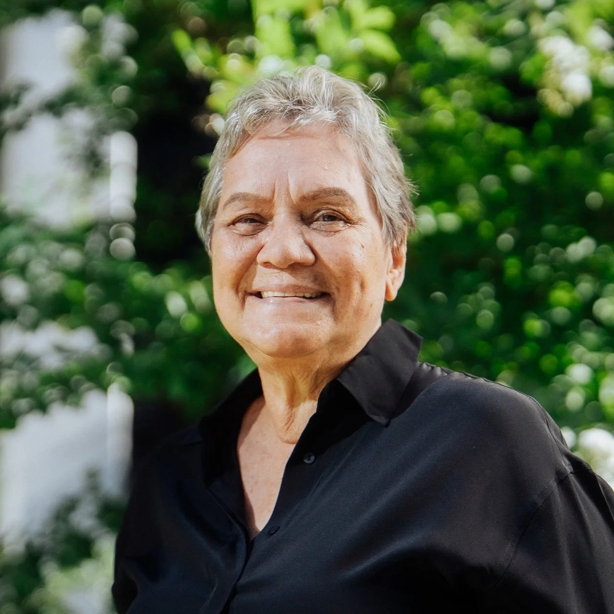 An elderly woman with short gray hair smiling outdoors with green foliage in the background, wearing a black shirt.