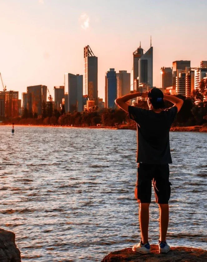 A person standing on a rock by a river, looking at a city skyline during sunset, with tall buildings and a partly cloudy sky.