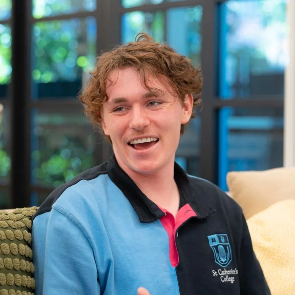 A young man with curly brown hair is smiling and talking, sitting on a beige couch in a bright room with large windows and greenery outside. He is wearing a colorful polo shirt with blue, black, pink, and red sections and a logo for St. Catherine's College.