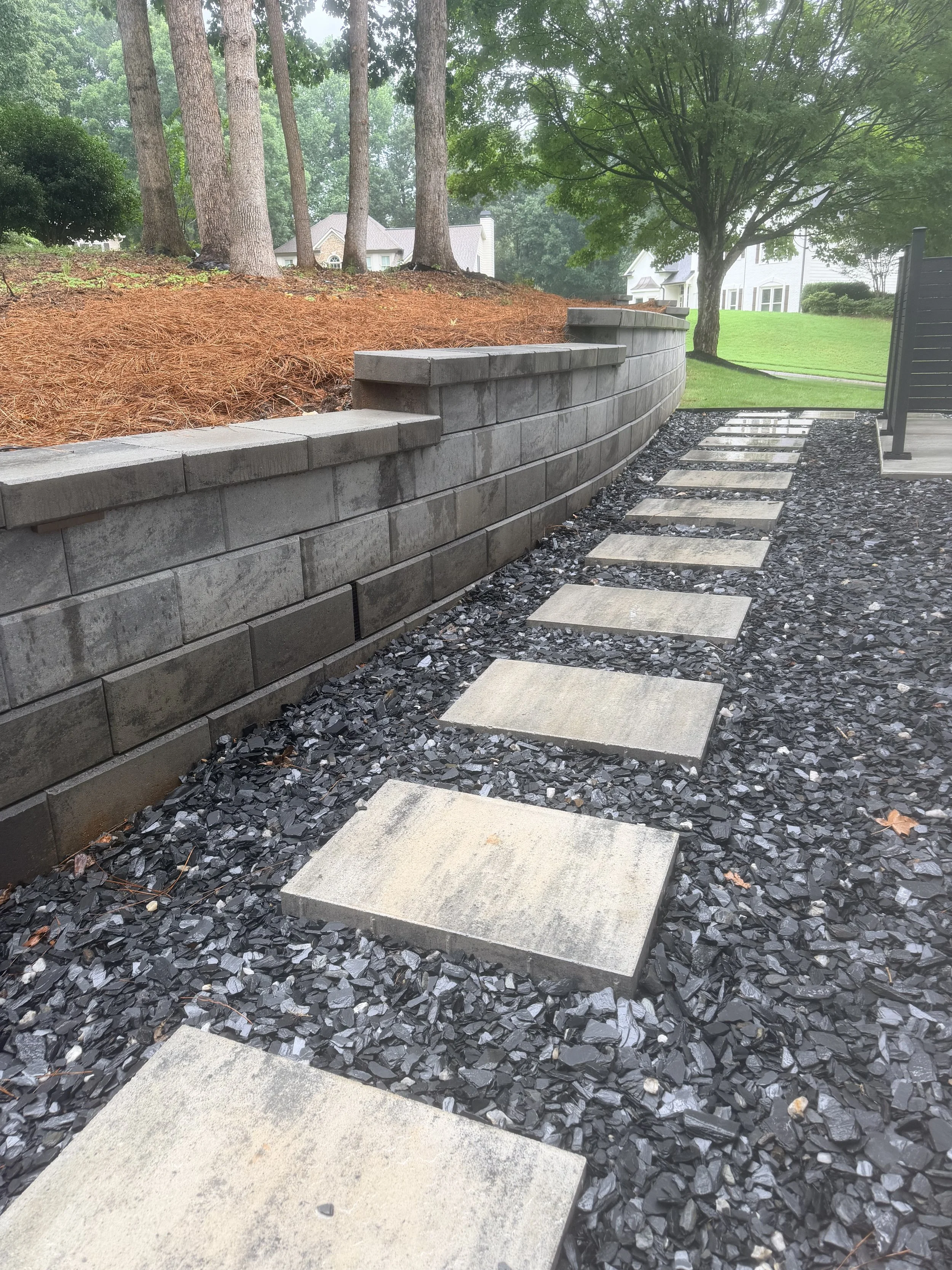 Concrete stepping stones on a gravel path next to a black metal fence, with a retaining wall made of concrete blocks on the left side, red mulch, trees, and a grassy yard in the background.