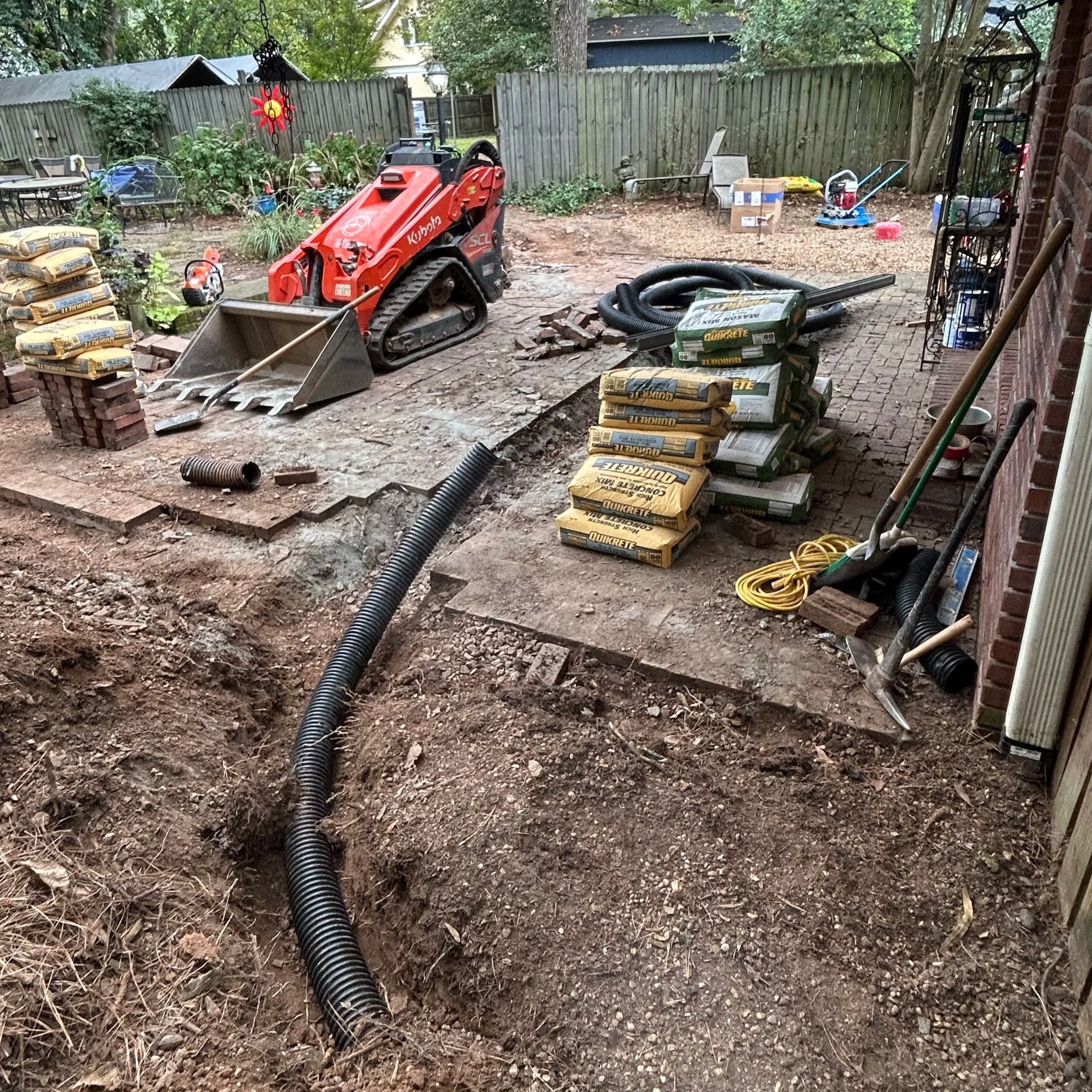 Backyard with construction work in progress, featuring a mini excavator, bags of concrete, hoses, and outdoor furniture.