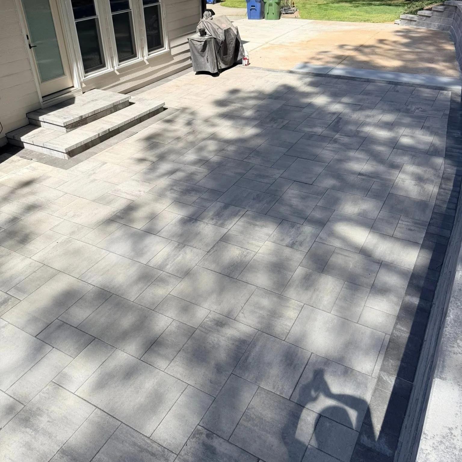 A backyard patio with large gray tiles, stairs leading to the door, and shadows from trees casting onto the surface.