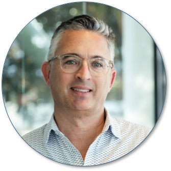 A smiling man with glasses and gray hair in a light-colored shirt, standing in front of a window with blurred outdoor scenery.