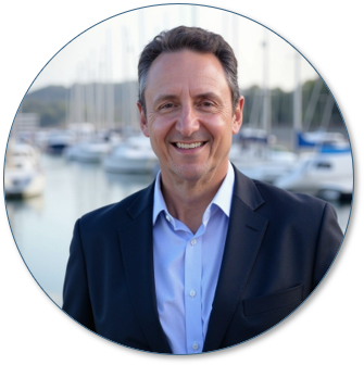 A smiling man in a suit standing outdoors near a marina with boats in the background.